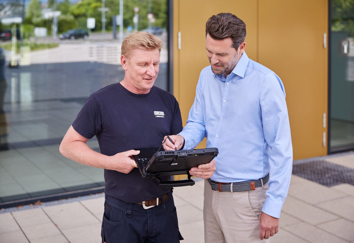 Service technician Michael Meyer explains something to a man wearing a shirt on his laptop in front of a building.