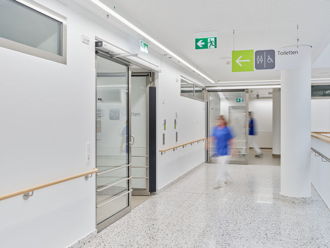 A nurse walks down a bright hospital corridor with sliding doors and signs for the toilets.