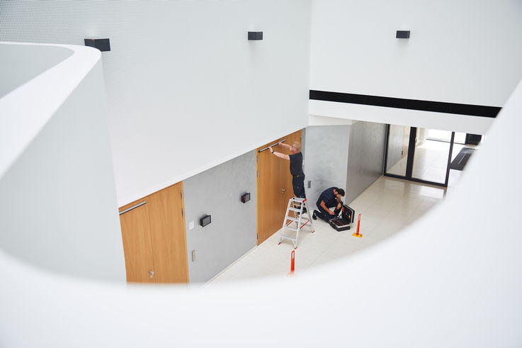 View from an open staircase to the lower floor, where two GEZE service technicians are installing a door closer on a door.