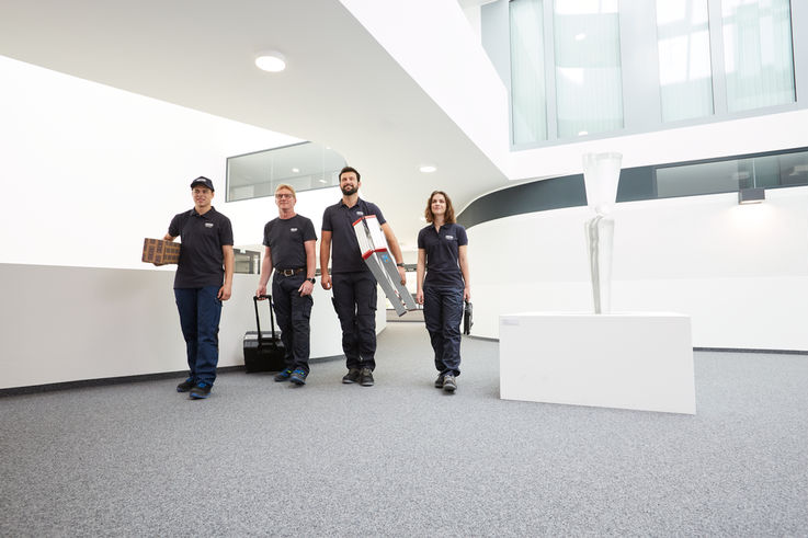 Four GEZE service technicians walk down a corridor in a modern building with their work materials.