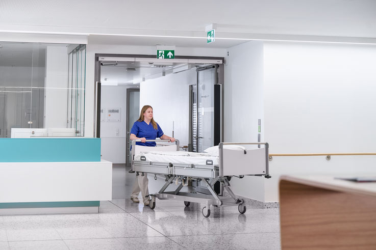 A nurse pushes an empty hospital bed through a door in the hospital corridor