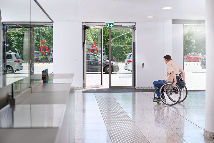 Man in wheelchair approaches an automatic glass door in a bright hallway