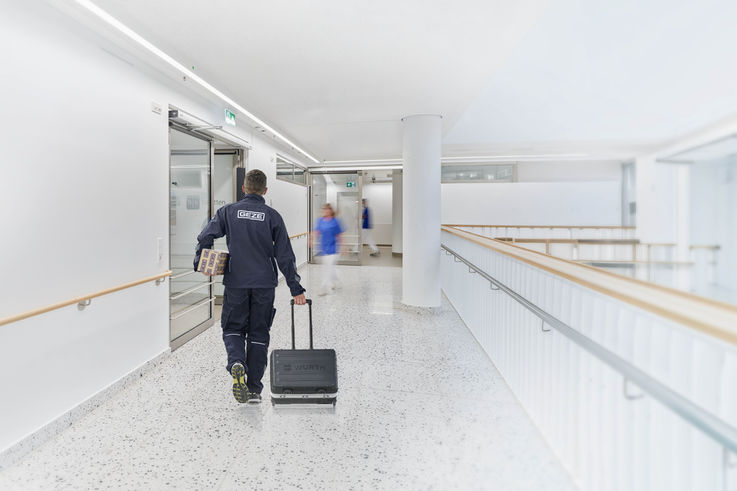 A GEZE service technician walks down a hospital corridor carrying a toolbox and a door closer